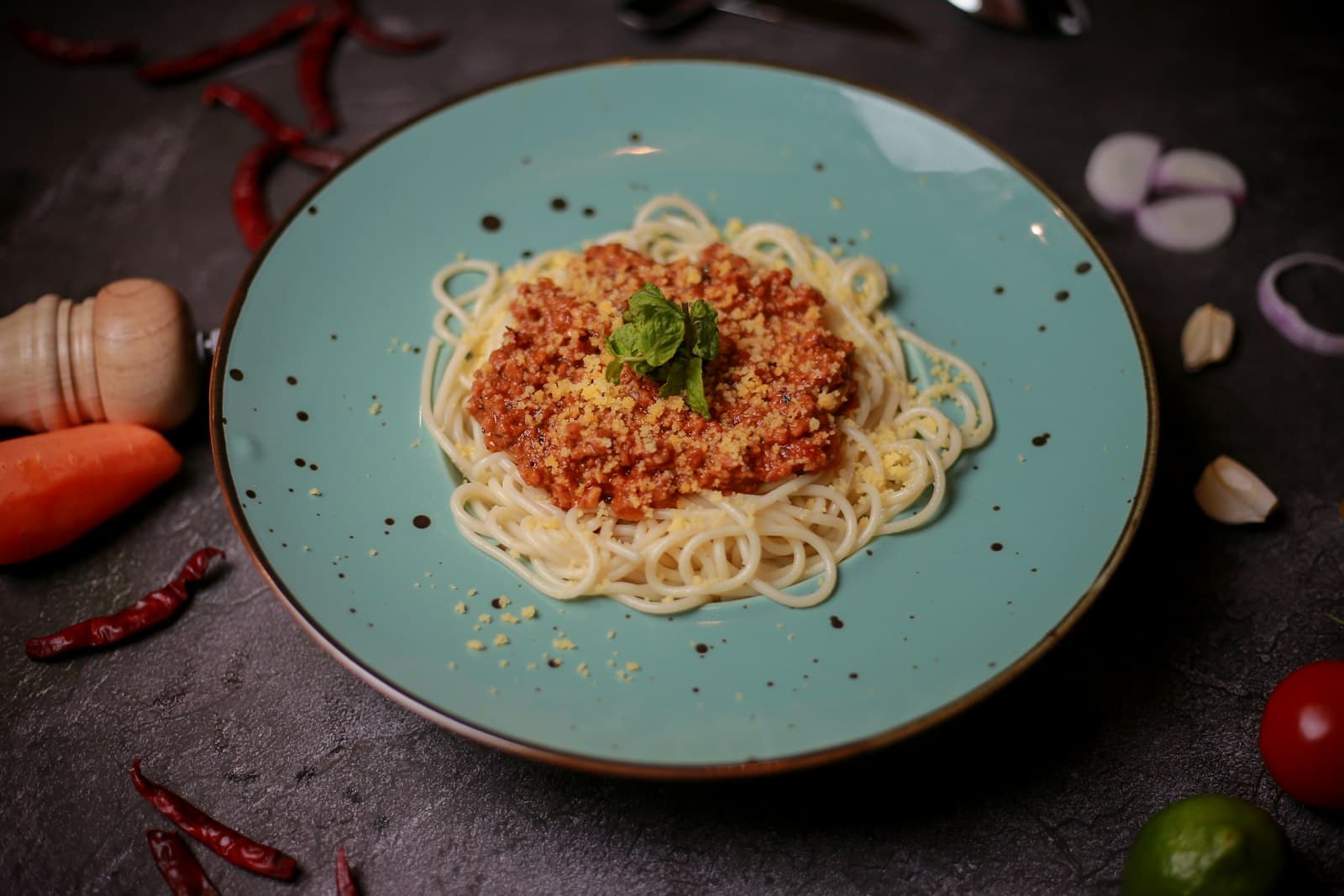 Spaghetti bolognese on a teal ceramic plate, dark moody background with chilis and herbs