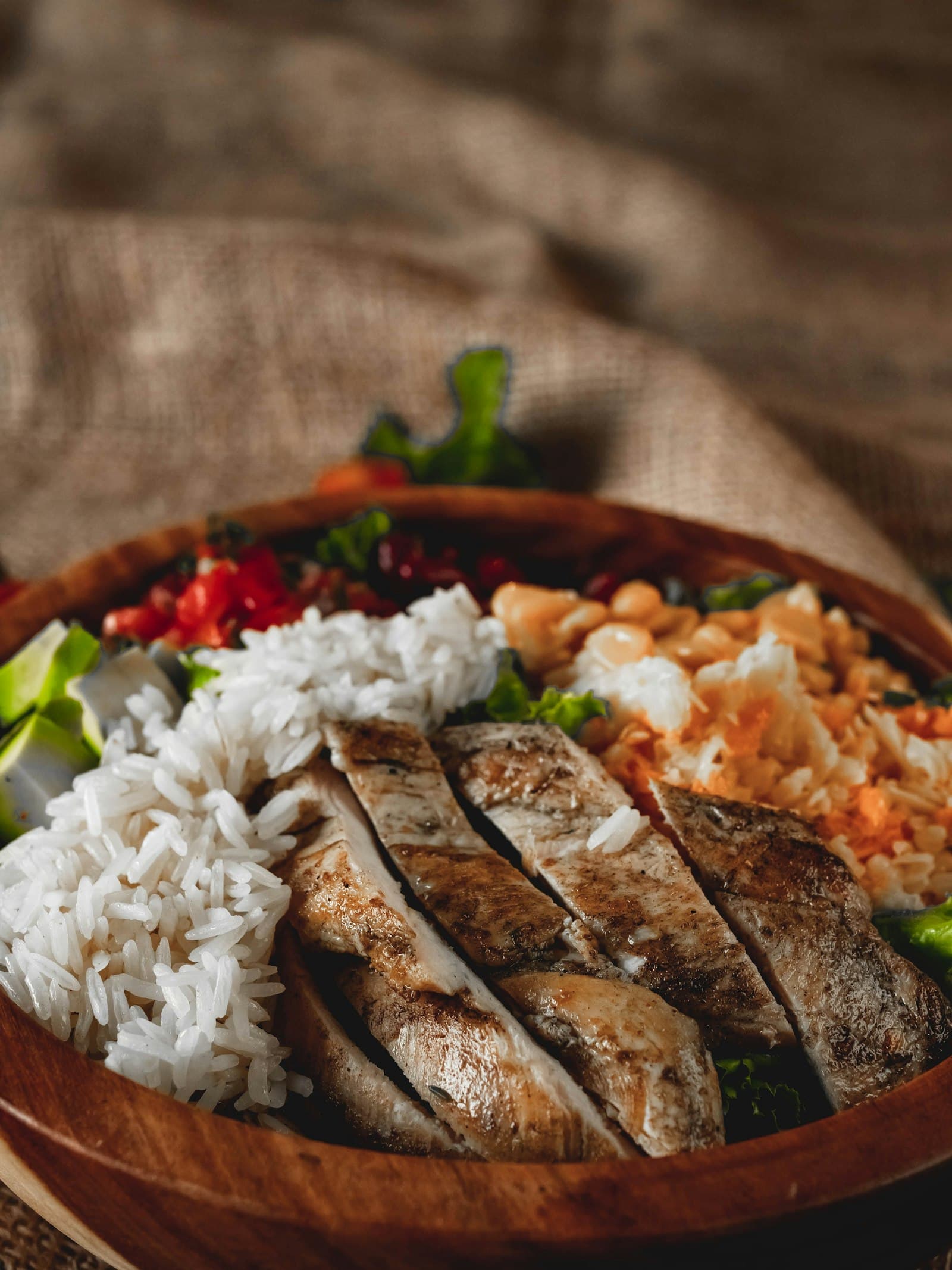Sliced grilled chicken breast over rice with peppers and broccoli in a wooden bowl
